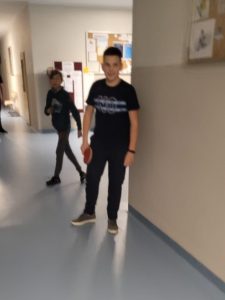 Uczeń stoi na korytarzu szkolnym z paletko do tenisa stołowego w dłoni. W tle widać innego ucznia. Zdjęcie lekko rozmazane.