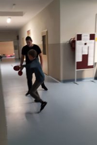 Dwóch uczniów klasy ósmej gra w tenisa stołowego na korytarzu szkolnym. Chłopcy w rękach trzymają paletki. Zdjęcie lekko niewyraźne.