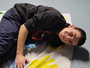 Uczeń klasy ósmej lezy na macie z namalowanymi kolorowymi rękami i stopami. Uczeń uśmiecha się do zdjęcia.