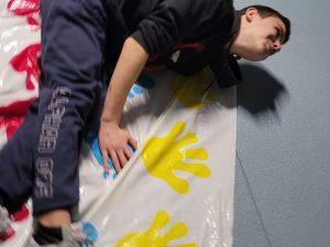 Uczeń klasy ósmej leży na maice z kolorowymi odciskami rąk i stóp. Chłopak wygina się na bok z uniesioną głową.