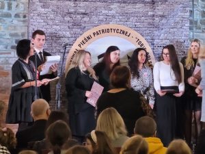 Na scenie stoją zebrane nauczycielki uczące nagrodzonych uczniów a wśród nich stoi Pani Iwona Szymańska. Po lewej stronie przy mikrofonach stoi dwoje osób prowadzących galę.