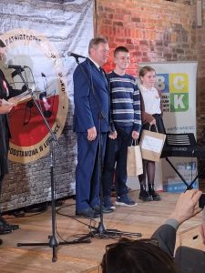 Na scenie stoi uczeń naszej szkoły nagrodzony w konkursie. Obok niego po prawej stoi inna nagrodzona uczestniczka konkursu a po lewej mężczyzna w granatowym garniturze.