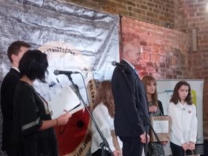 Na scenie stoją Państwo przemawiający do mikrofonu oraz nagrodzeni uczestnicy konkursu. Po prawej stronie z przefu stoi uczennica naszej szkoły.