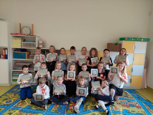 Uczniowie klasy I pozują do grupowego zdjęcia w swojej sali. Dzieci trzymają w dłoniach przed sobą identyczne książki. Za dziećmi widać kremową ścianę i stojącą przed nią meblościankę.