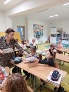 Uczniowie klasy pierwszej siedzą przy stolikach. Wśród nich chodzi Pani Sylwie - bibliotekarka trzymająca w dłoniach stos książek. Pani podaje jednej z uczennic książkę.