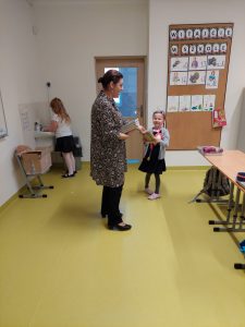 Zdjęcie stojącej w sali Panibibliotekarki trzymającej stos ksiązek. Pani podaje uczennicy klasy pierwszej jedną z książek. W tle widać stojącą przy umywalce inną uczennicę.