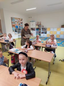 Uczniowie klasy pierwszej siedzą przy stolikach. Wśród nich chodzi Pani Sylwie - bibliotekarka trzymająca w dłoniach stos książek.