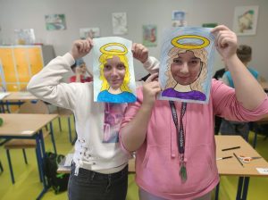 Dwie uczennice przykładają do twarzy wykonane z papieru ramki z narysowanymi włosami i aureolą nad nimi. Dziewczynki stoją obok siebie. W tle widać salę lekcyjną.