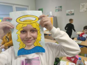 Uczennica przykłada do twarzy wykonaną z papieru ramkę z narysowanymi włosami i aureolą nad nimi. Dziewczynka się usmiecha.