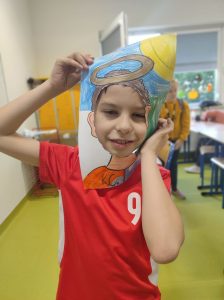 Uczeń przykłada do twarzy wykonaną z papieru ramkę z narysowanymi krótkimi włosami i aureolą nad nimi. Chłopiec lekko przyciska ramke do twarzy z prawej strony.