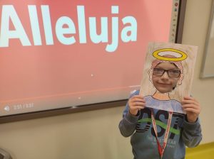 Uczeń przykłada do twarzy wykonaną z papieru ramkę z narysowanymi krótki włosami i aureolą nad nimi.