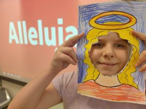 Uczennica przykłada do twarzy wykonaną z papieru ramkę z narysowanymi włosami i aureolą nad nimi. W tle widać wyświetlający się fragment napisu ALLELUJA