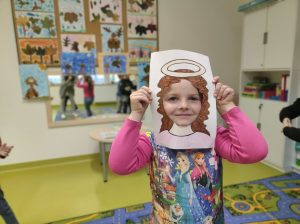 Uczennica przykłada do twarzy wykonaną z papieru ramkę z narysowanymi włosami i aureolą nad nimi.