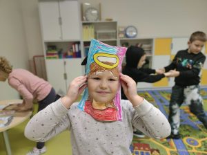 Uczennica przykłada do twarzy wykonaną z papieru ramkę z narysowanymi włosami i aureolą nad nimi. W tle widać bawiące się dzieci.