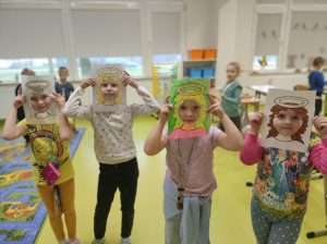 Cztery uczennice przykładają do twarzy wykonane z papieru ramki z narysowanymi włosami i aureolą nad nimi. Dziewczynki stoją w jednym rzędzie a za nimi widać salę lekcyjną.