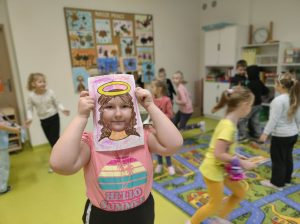 Uczennica przykłada do twarzy wykonaną z papieru ramkę z narysowanymi włosami i aureolą nad nimi. W tle widać biegające dzieci.