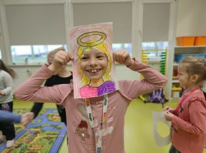 Uczennica przykłada do twarzy wykonaną z papieru ramkę z narysowanymi włosami i aureolą nad nimi. Dziewczynka szeroko się usmiecha.