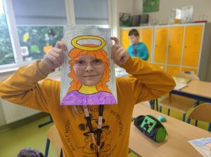 Uczennica przykłada do twarzy wykonaną z papieru ramkę z narysowanymi włosami i aureolą nad nimi.