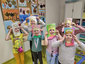 Sześć uczennic stojących obok siebie pozuje do zdjęcia. Dziewczynki trzymają w dloniach papierowe ramki z narysowanymi włosami i aureolą nad nimi.