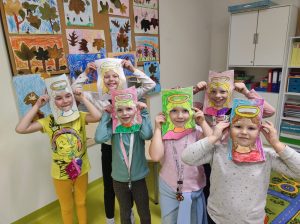 Sześć uczennic stojących obok siebie pozuje do zdjęcia. Dziewczynki trzymają w dloniach papierowe ramki z narysowanymi włosami i aureolą nad nimi. Dziewczynki się uśmiechają.