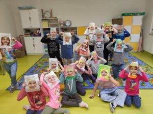 Uczniowie klasy pierwszej pozują do wspólnego zdjęcia na dywanie. Wszyscy przy twarzach trzymają wykonane z papieru ramki z włosami i aureolą nad nimi.