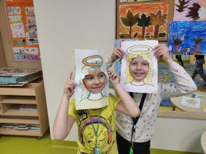 Dwie uczennice przykładają do twarzy wykonane z papieru ramki z narysowanymi włosami i aureolą nad nimi. Dziewczynki trzymają ramki w podniesiono wysoko w góre rękach i stoją blisko obok siebie.