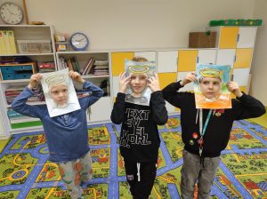 Trzech uczniów przykłada do twarzy wykonane z papieru ramki z narysowanymi krótkimi włosami i aureolą nad nimi. Chłopcy stoją w jednym rzędzie obok siebie. W tle widać dywan i meblościankę ustawioną pod scianą sali lekcyjnej.