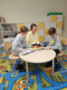 Trzy uczennice klasy piątej siedzą przy okrągłym stoliku i oglądają leżące na stoliku otwarte książki.