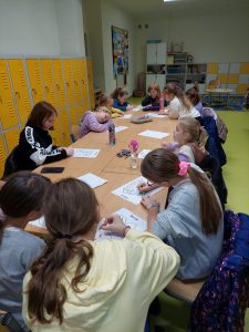 Uczniowie siedzą przy długim wspólnym stole utworzonym ze szkolnych ławek. Uczniowie kolorują wydrukowane rysunki.