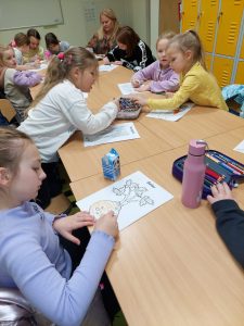 Uczniowie siedzą przy długim wspólnym stole utworzonym ze szkolnych ławek. Uczniowie kolorują wydrukowane rysunki.