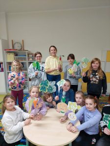 Uczniowie klasy pierwszej wraz z trzema uczennicami klasy piątej pozują do wspólnego zdjęcia. Część dzieci siedzi przy okrągłym stoliku. Wszystkie dzieci trzymają patyczki z przyklejonymi obrazkami warzyw, które wcześniej kolorowały.