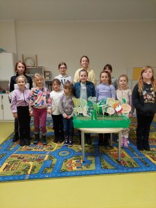 Uczennice klasy pierwszej i trzy uczennice klasy piatej pozują do wspólnego zdjęcia. Dziewczynki stoją w dwóch rzędach a przed nimi na okragłym stole ustawiono makietę z ustawionymi patyczkami z rysunkami warzyw.