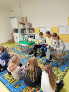 Uczennice klasy piątej siedzą obok siebie na krzesełkach. Dziewczynki mają na kolonach rozłożone książki. Przed nimi siedzą ich młodsze koleżanki a obok stoi stoli z makietą warzyw.