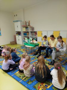 Uczennice klasy piątej siedzą obok siebie na krzesełkach. Dziewczynki mają na kolonach rozłożone książki. Przed nimi siedzą ich młodsze koleżanki a obok stoi stoli z makietą warzyw.