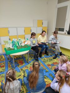 Trzy uczennice klasy piętej siedzą obok siebie na krzesełkach. Obok nich na stoliku stoi makieta z kolorowymi warzywami narysowanymi na kartkach. Na przeciwko uczennic siedzą uczniowie lasy pierwszej.