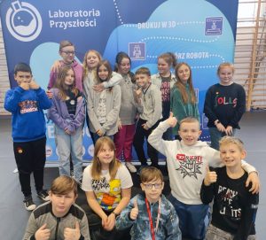 Uczniowie klasy czwartej pozują do grupowego zdjęcia na tle niebieskiego baneru z napisem Laboratorium Przyszłości. Część uczniów podnosi w górę kciuki.