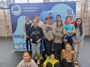 Uczniowie klasy szóstej pozują do grupowego zdjęcia na tle niebieskiego baneru z napisem Laboratorium przyszłości. Troje uczniów siedzi na podłodze, reszta stoi. Jeden z uczniów siedzących na podłodze opiera się o stopy stojących za nim kolegów.