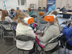 Dwie uczennice z goglami VR na głowie odracają się do tyłu. Jedna z dziewczynek trzyma w dłoni czarną kostkę z białymi rysunkami. Przed dziewczynkami siedzą na krzesełkach inni uczniowie.