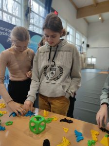 Dwie uczennice klasy szóstej stoją przy stoliku i oglądają przedmioty wydrukowane w drukarce 3D. Dziewczynki dotykają dłoniami wybranych przez siebie przedmiotów.