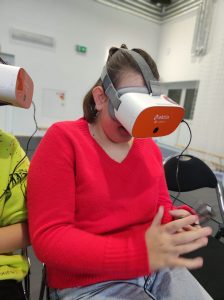 Uczennica klasy szóstej siedzi na krzesełku z założonymi na głowie goglami VR. Dziewczynka ogląda przez nie wyciągnięte przed siebie dłonie.