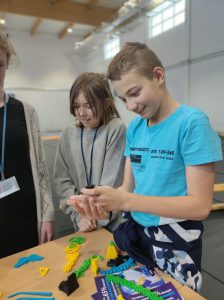 Dwoe uczniów klasy szóstej stoi przy stoliku z ułożonymi przedmiotami wydrukowanymi na drukarce 3D. Uczeń trzyma w dloniach przed sobą jeden z przedmiotów i się uśmiecha.