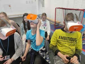 Troje uczniów klasy szóstej siedzi obok siebie na krzesełkach z goglami VR na głowie. Jeden z chłopców spogląda wysoko w górę. Pozostała dwójka patrzy przed siebie.