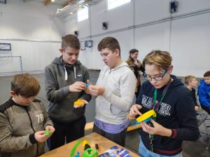 Czworo uczniów stoi przy stoliku z przedmiotami wydrukowanymi w drukarce 3D. Chłopcy oglądają trzymające w dłoniach przedmioty.