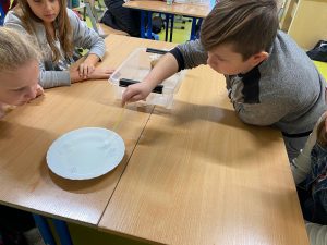 Uczeń klasy czwartej trzymający pipetkę w dłoni pochyla się nad stolikiem z białym talerzem wypełnionym wodą. Na przeciwko chłopca siedzą dwie dziewczynki przyglądające się wykonywanej przez chłopca czynności.