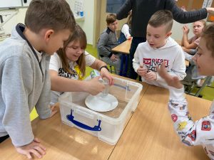 Uczniowie klasy czwartej stoją nad plastikowym pudełkiem zumieszczonym w środku bialym talerzem. Dziewczynka pochylająca się nad pudelkiem przelewa wodę z plastikowego kubka do szklanego naczynia. Troje uczniów stojących obok przygląda się jej z zainteresowaniem.