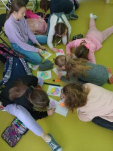Na podłodze siedzą uczniowie w grupce. Przed nimi leżą rysunki diagramów kołowych oraz piórniki z kolorowymi kredkami i flamastrami. Zdjęcie wykonane w pewnym oddaleniu.
