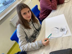 Uczennica siedzi w ławce z ołówkiem w ręku. Przed nia leży kartka z rysunkiem. Dziewczynka spogląda i lekko usmiecha się w kierunku aparatu.