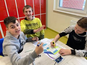 Trzech uczniów siedzi przy wspólnym stole i tworzy plakat. Dwóch spogląda w stronę aparatu a trzeci z opuszczoną głową koloruje na żółto rysunek domu.