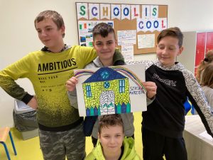 Czworo uczniów pozuje do zdjęcia ze swoim plakatem zatytułowanym SZKOŁA MARZEŃ. Trzech chłopców stoi a czwarty przed nimi kuca. Tuż nad jego głową stojący po srodku uczeń trzyma plakat przedstawiający budynek i wychodzącą z niego tęczę.