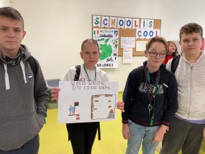 Czworo uczniów stoi obok siebie. Dziewczynka stojąca jako druga od lewej strony trzyma w dłoniach przed sobą plakat zatytułowany: WARTO WALCZYC O TO CO SIĘ KOCHA. Na rysunku domek w ramce oraz postać trzymająca flagę.